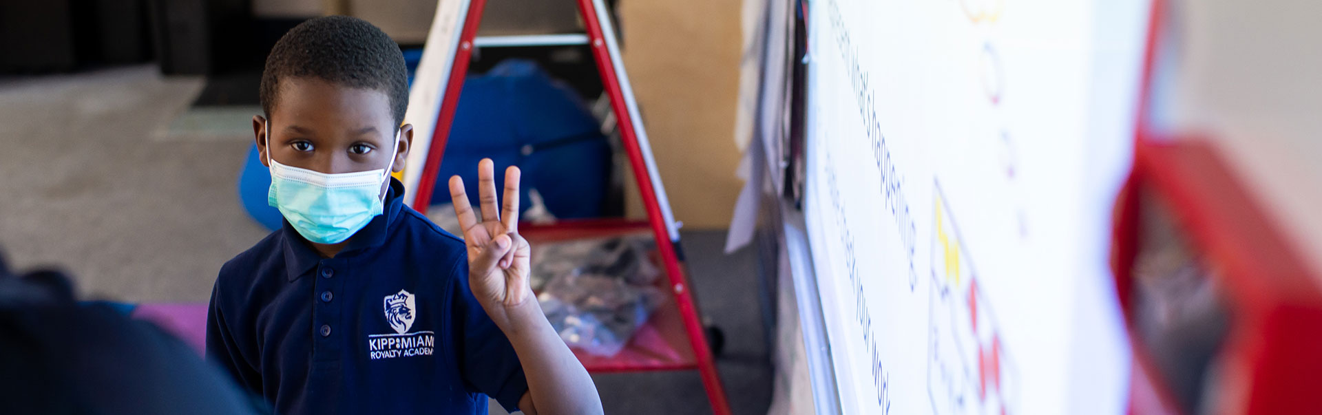 young student in classroom waving to camera